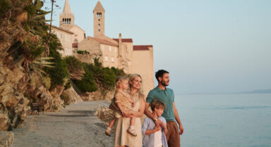 Two adults and two children stand on a seaside promenade near stone buildings and church towers of Rab town, looking out over calm water.