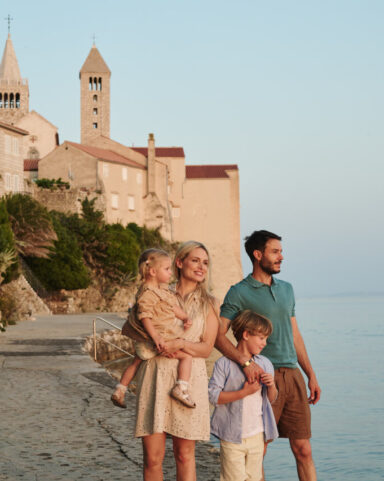 Two adults and two children stand on a seaside promenade near stone buildings and church towers of Rab town, looking out over calm water.