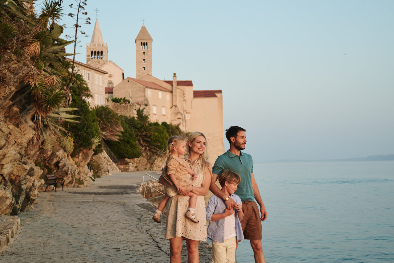 Two adults and two children stand on a seaside promenade near stone buildings and church towers of Rab town, looking out over calm water.