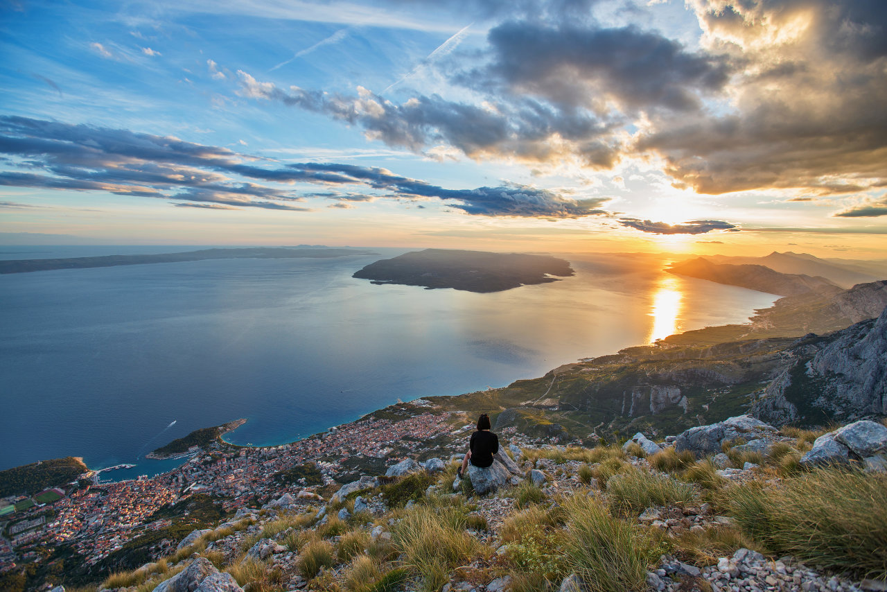A person sits on a rocky mountainside overlooking the Adriatic and Makarska at sunset. 