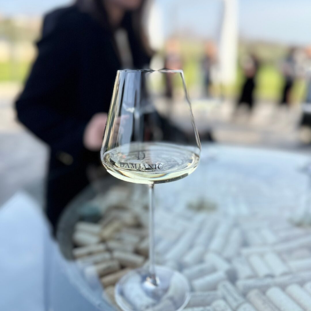 A glass of Virtu Brut with the Damjanić logo, served on a table decorated with wine corks, with vineyards softly blurred in the background. A glass of Virtu Brut with the Damjanić logo, served on a table decorated with wine corks, with vineyards softly blurred in the background.