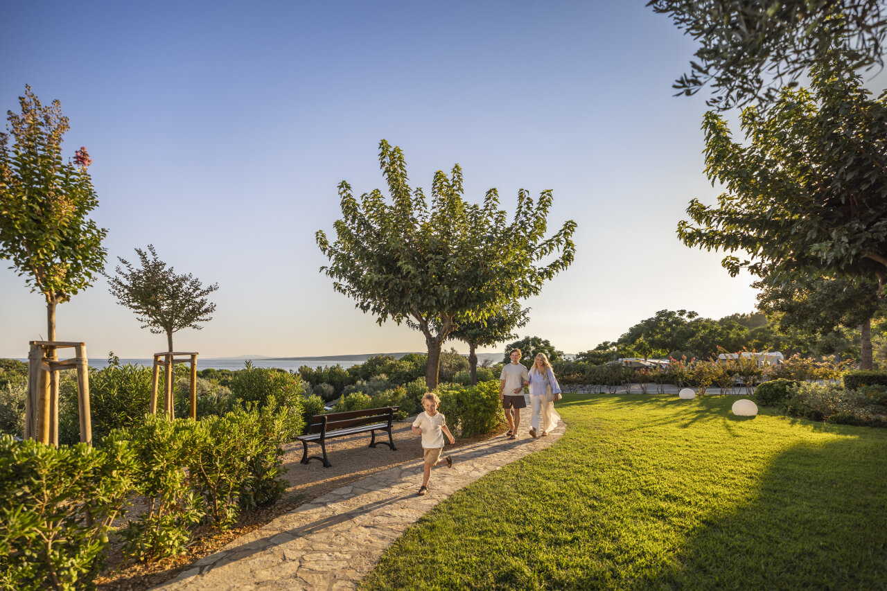 A child runs ahead of two adults along a curved stone path through a sunny garden with trees, shrubs, and sea views in the distance.