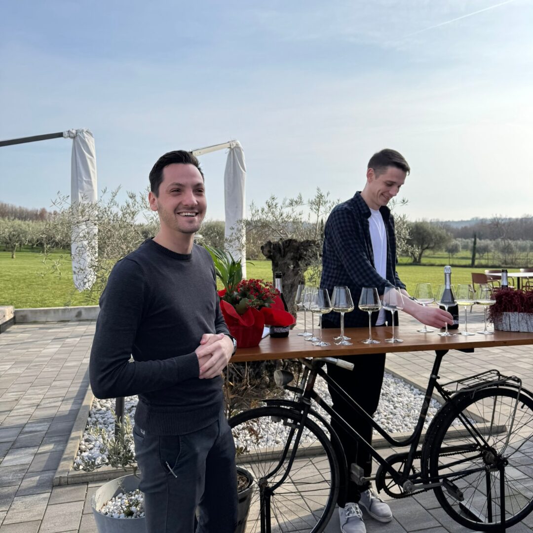 Two hosts preparing a wine tasting on an outdoor terrace surrounded by vineyards, with wine glasses arranged on a wooden table set on a bicycle stand. Two hosts preparing a wine tasting on an outdoor terrace surrounded by vineyards, with wine glasses arranged on a wooden table set on a bicycle stand.
