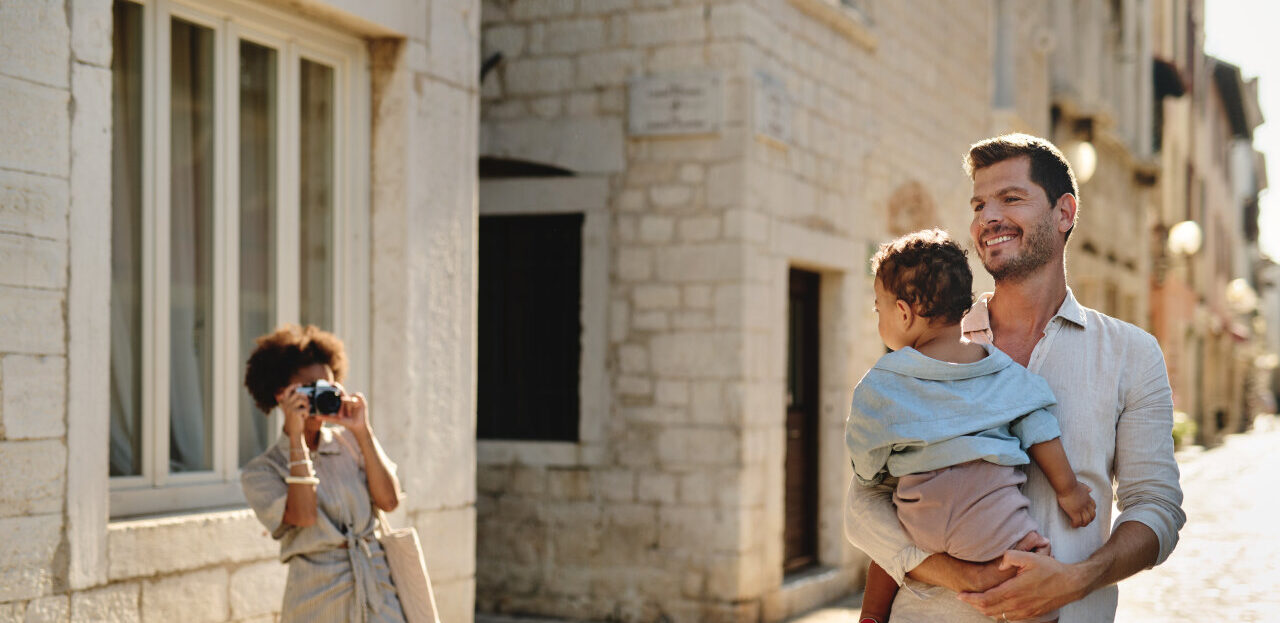 Ein Vater h&auml;lt ein Kleinkind im Arm und eine Frau fotografiert sie in einer sonnenbeschienenen Steingasse in der Altstadt von Poreč. 