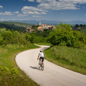 A cyclist riding along a winding road through green hills towards a hilltop village in the Istrian countryside.