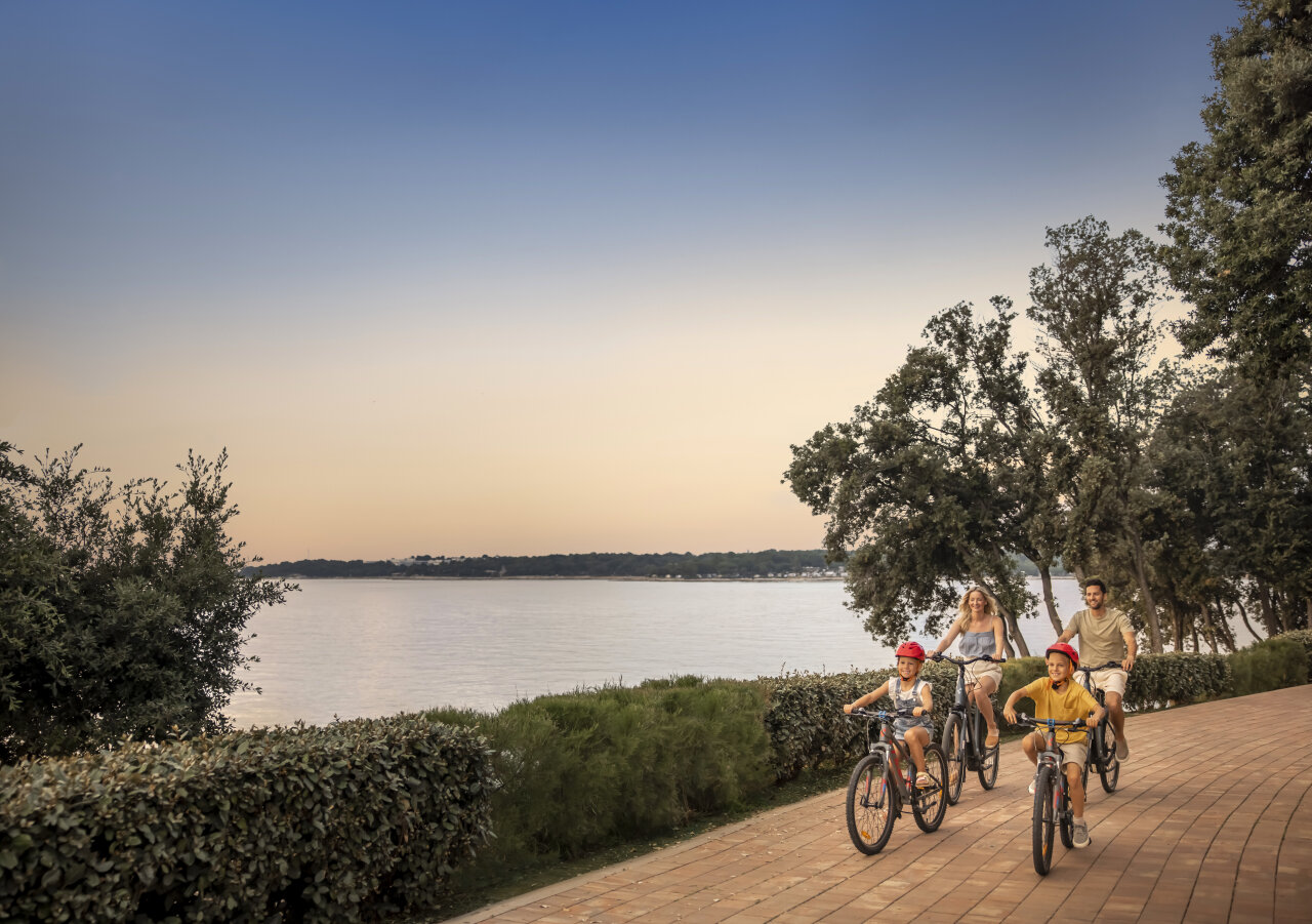 A family with two children cycling along a landscaped seafront promenade at sunset in Valamar Camping Istra.