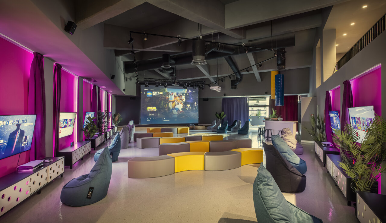 A spacious indoor gaming zone with a large screen, consoles, bean bags and ambient pink lighting. 